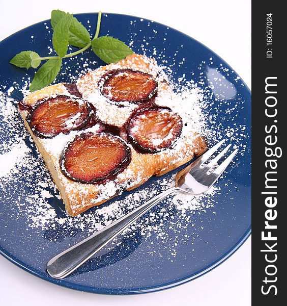 Piece of Plum Pie covered with powder sugar on a blue plate decorated with a mint twig