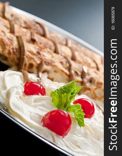 Three rolled pancakes close-up arranged with cream, cherries, chocolate and sprig mint leaves in black ceramic plate. Three rolled pancakes close-up arranged with cream, cherries, chocolate and sprig mint leaves in black ceramic plate