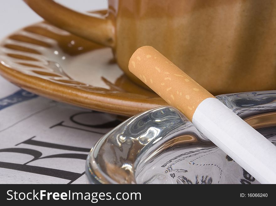 A filtered cigarette resting on a glass ashtray next to a brown cup and plate and a newspaper. A filtered cigarette resting on a glass ashtray next to a brown cup and plate and a newspaper.