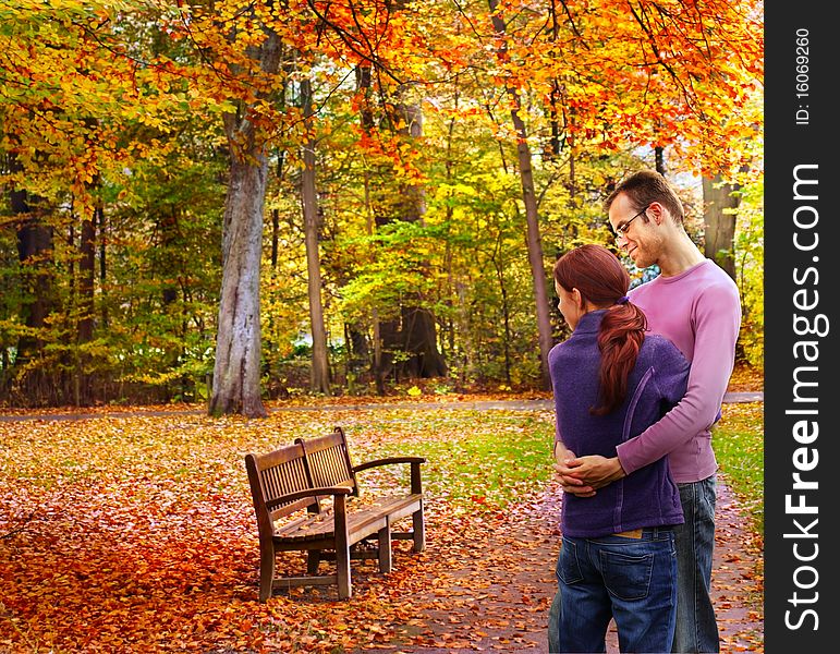 Happy couple standing embraced in the autumnal park looking to a bench. Happy couple standing embraced in the autumnal park looking to a bench
