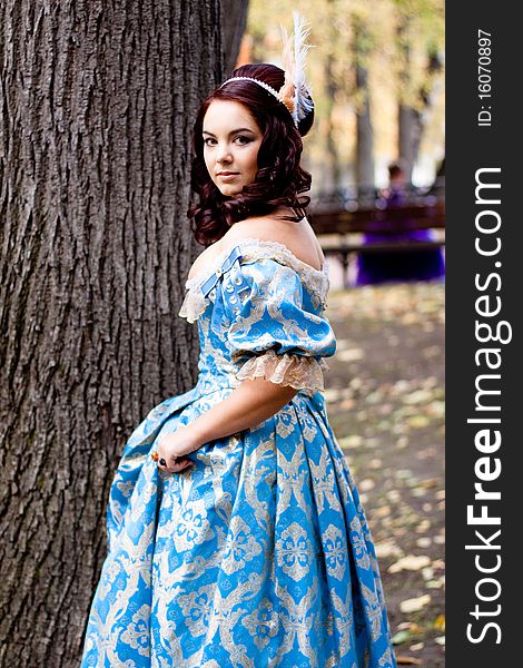 A portrait of lady in a blue baroque dress standing near a bole. A portrait of lady in a blue baroque dress standing near a bole