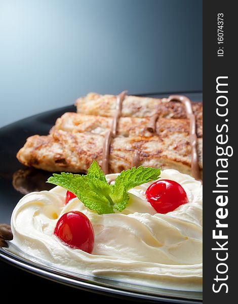 Three rolled pancakes close-up arranged with cream, cherries, chocolate and sprig mint leaves in black ceramic plate. Three rolled pancakes close-up arranged with cream, cherries, chocolate and sprig mint leaves in black ceramic plate