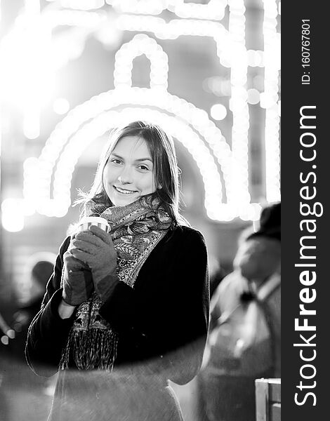 Black and white photo of young girl with glass in her hands on street in evening against background of burning garlands