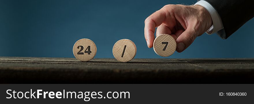 Wide view image of a hand of a businessman assembling a 24/7 sign with wooden cut circles. Over blue background. Wide view image of a hand of a businessman assembling a 24/7 sign with wooden cut circles. Over blue background