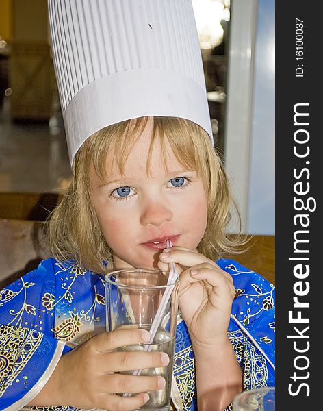 Little beautiful girl in blue dress and cook hat drinking water in stow. Little beautiful girl in blue dress and cook hat drinking water in stow.