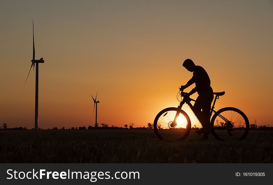 A cyclist with a camera stands in a field during the sunset against the background of an orange sky with the sun and windmills