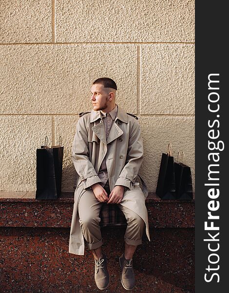 A stylish young man is wearing a beige trench coat and posing next to the old building. He is holding bags after successful shopping on Black Friday. A stylish young man is wearing a beige trench coat and posing next to the old building. He is holding bags after successful shopping on Black Friday