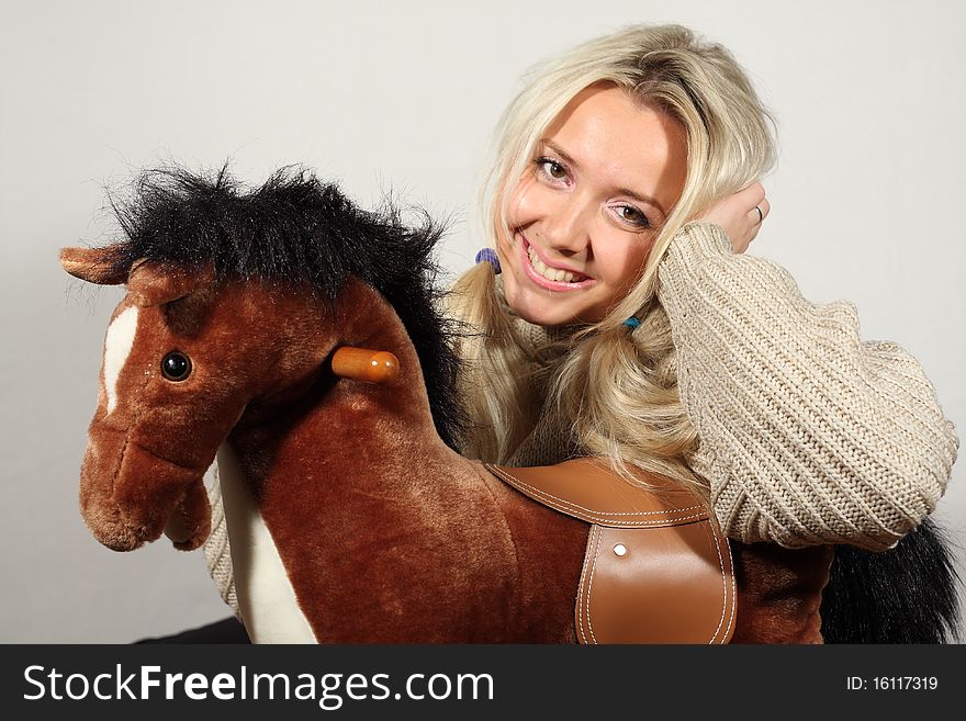Toy horse and blond hair lady weared winter sweat shot, smiles by gray background. Toy horse and blond hair lady weared winter sweat shot, smiles by gray background.