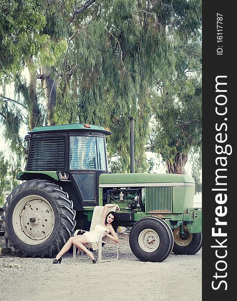 Country farm girl, beautiful young woman wearing a dress sitting on chair beside tractor