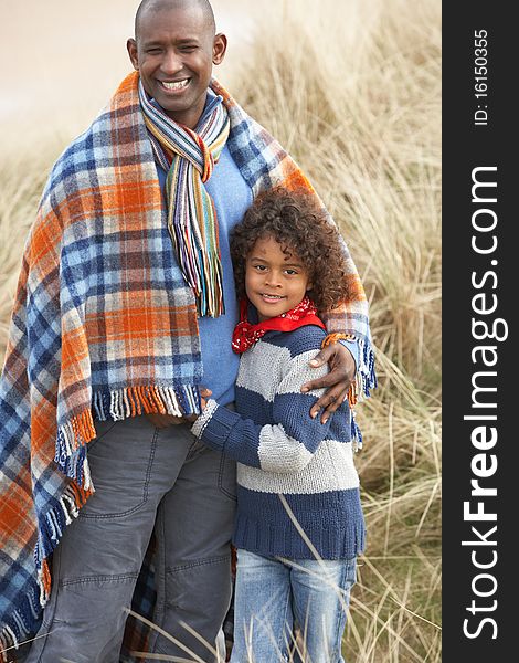 Father And Son Wrapped In Blanket Amongst Sand Dunes On Winter Beach Smiling. Father And Son Wrapped In Blanket Amongst Sand Dunes On Winter Beach Smiling