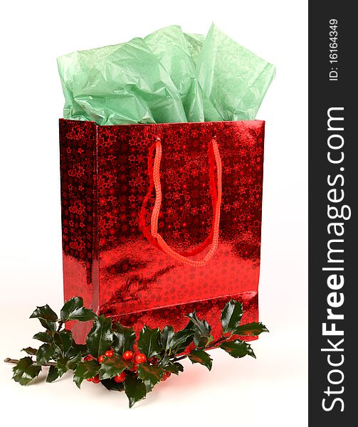 Red shiny Christmas gift bag with green tissue and a sprig of holly with red berries on white with soft shadow. Red shiny Christmas gift bag with green tissue and a sprig of holly with red berries on white with soft shadow.