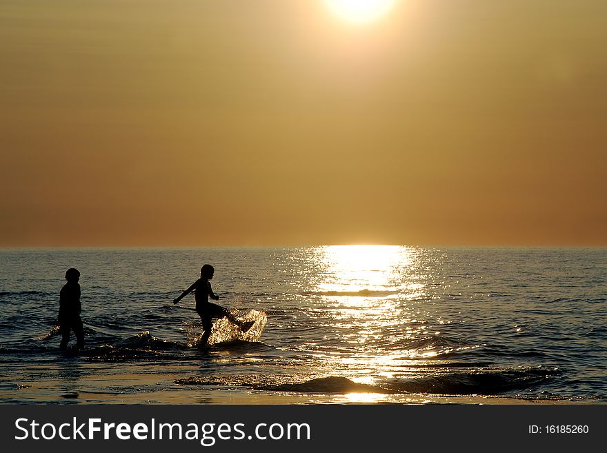 Two silhouetted kids kick,play and splash in the sparkling reflection on the blue rippled water, hazy orange sky by half sunlight, warm colors, soft waves, heat from sun and cool splashing of water.