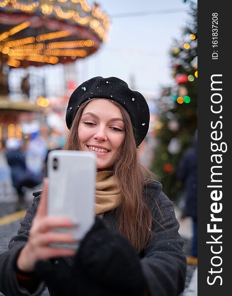 Beautiful young woman taking a selfie with christmas tree behind her with mobile phone camera - Pretty girl enjoying