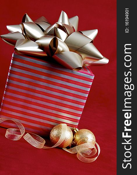 A package wrapped in silver and red paper is adorned with a large silver bow. Gold frosted ornaments and transparant gold ribbon rest in front of the package. All objects are sitting atop a red velvet background. A package wrapped in silver and red paper is adorned with a large silver bow. Gold frosted ornaments and transparant gold ribbon rest in front of the package. All objects are sitting atop a red velvet background.