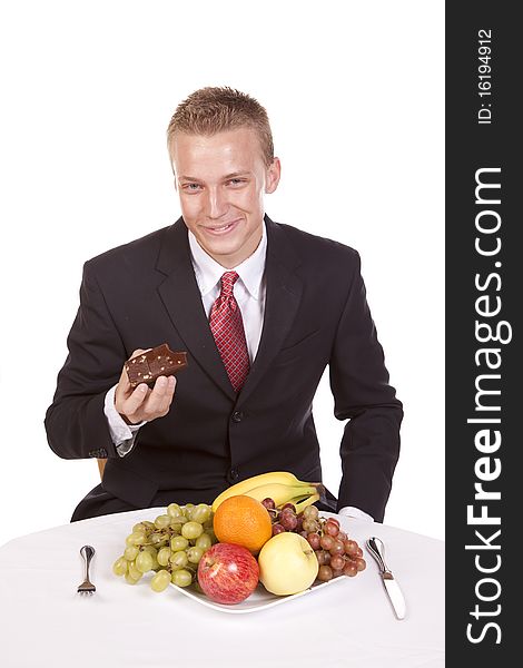 A man deciding to eat a brownie instead of his fruit on a plate in front of him. A man deciding to eat a brownie instead of his fruit on a plate in front of him.