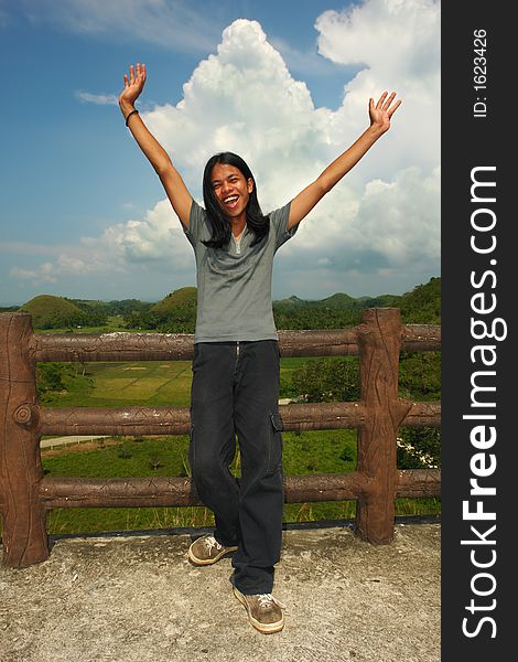 Asian exotic long-haired boy cheering, hands up, in front of the touristy landmark of the Chocolate Hills in Bohol, Philippines. Asian exotic long-haired boy cheering, hands up, in front of the touristy landmark of the Chocolate Hills in Bohol, Philippines.