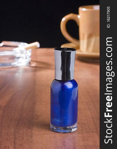Blue nail polish standing on a coffee table in front of a cup of coffee and an ashtray.