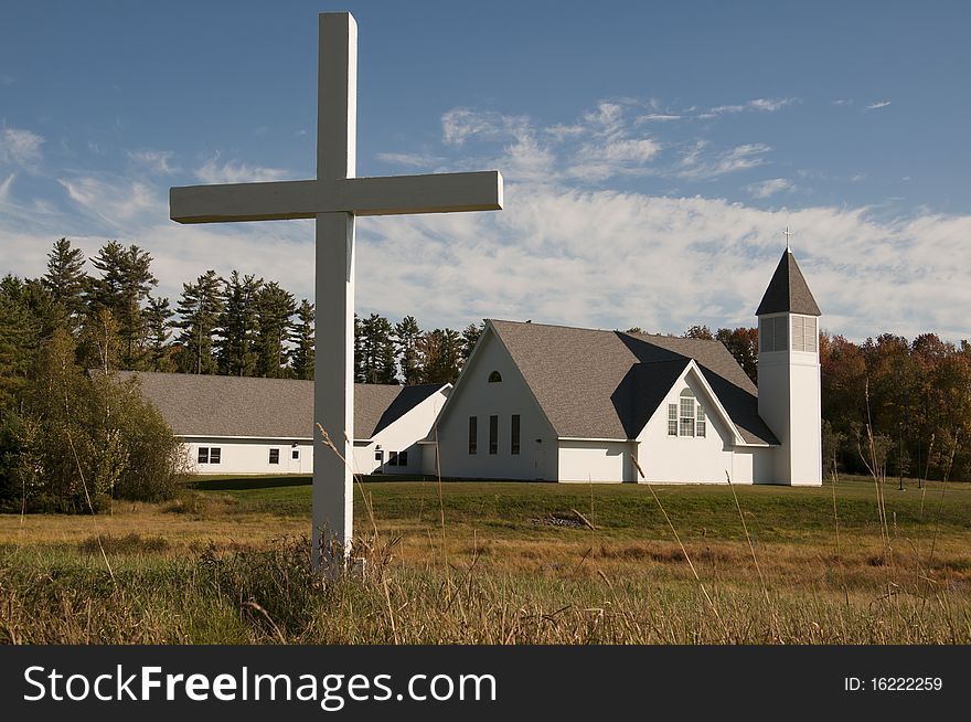 A single white cross stands in front of a simple white church in a rural setting. A single white cross stands in front of a simple white church in a rural setting.