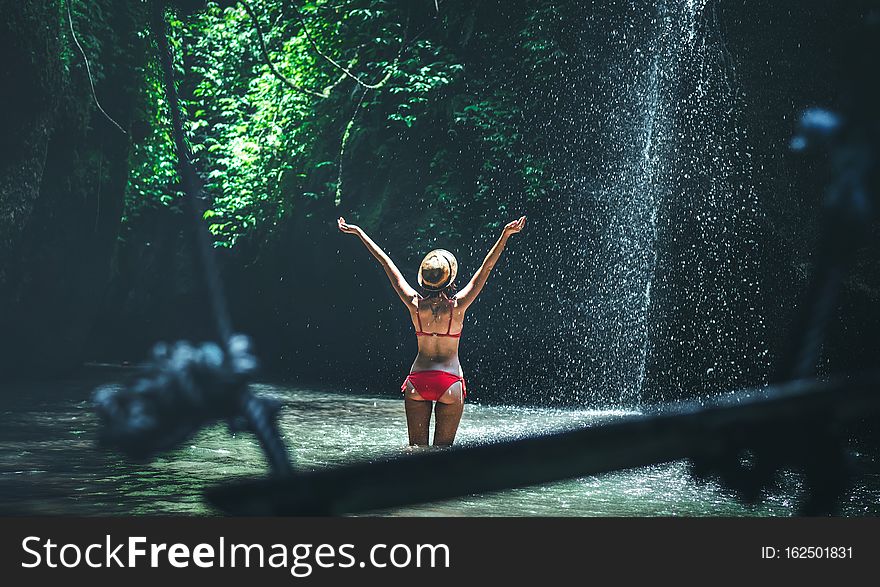 Young woman tourist with straw hat deep in the rainforest with waterfall background. Bali island. Indonesia.