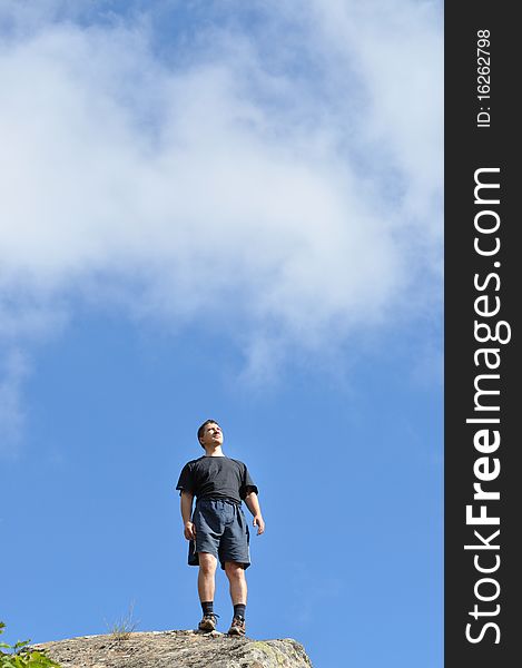 A young man stands on a cliff top, and seriously, confidently looking toward the background blue sky