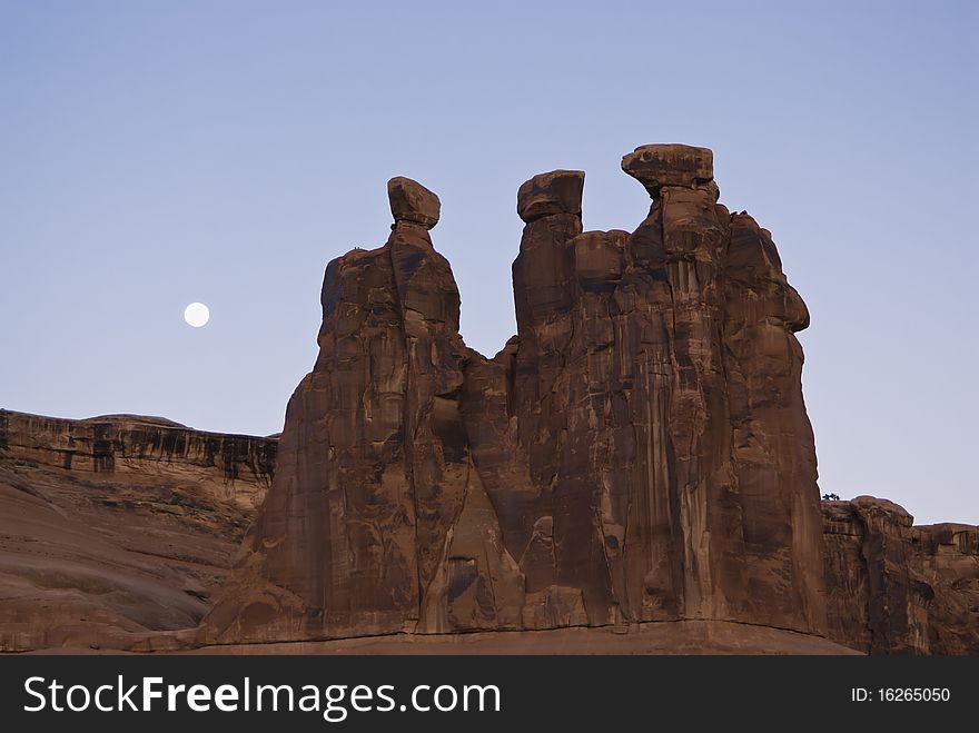 A full moon sets behind the Three Gossips formation in Arches National Park.