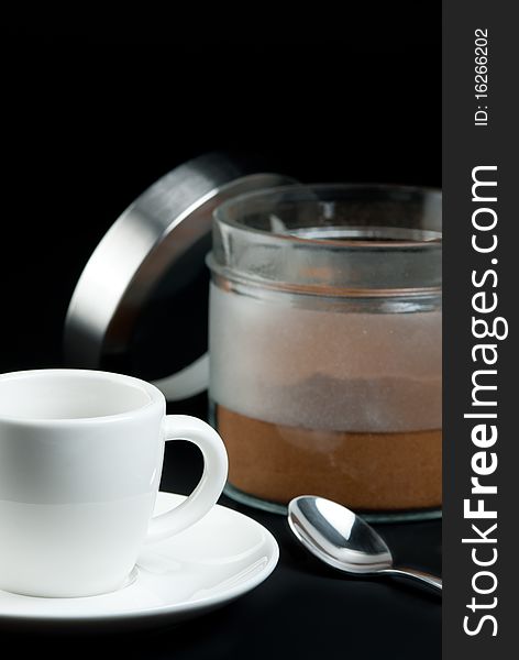 Coffee preparing - White cup on saucer with tea spoon and open dose with ground coffee on black background.