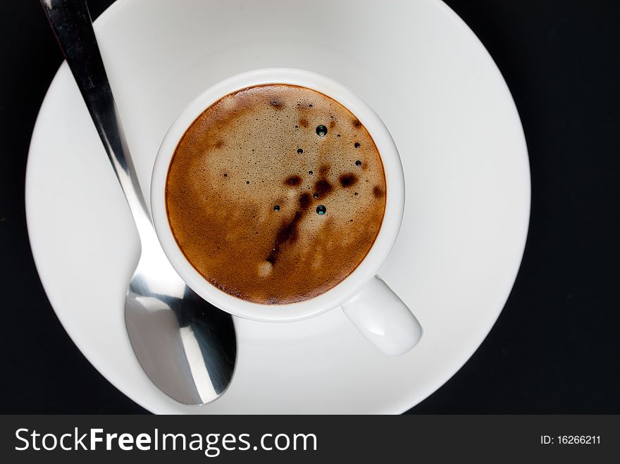 Top view of coffee with mousse in white cup standing on saucer with tea spoon on black background. Top view of coffee with mousse in white cup standing on saucer with tea spoon on black background.