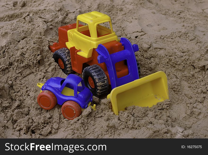 The plastic toy tractor with the small blue car on the sandbox. The plastic toy tractor with the small blue car on the sandbox