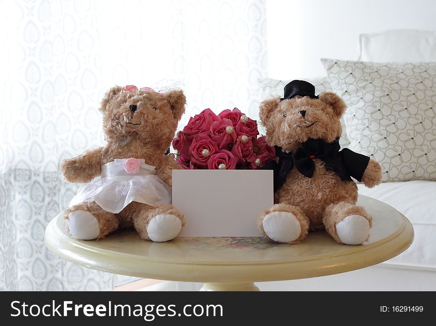 A pair of cute bride and groom teddy bears with a blank note suitable for wedding theme with a bouquet of pink roses at the back. A pair of cute bride and groom teddy bears with a blank note suitable for wedding theme with a bouquet of pink roses at the back