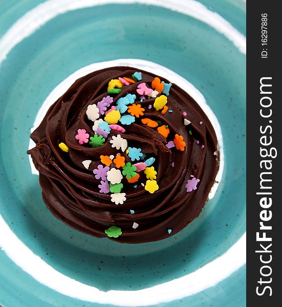 A delicious chocolate frosted cupcake with multicolored sprinkles sitting on a bright blue plate as seen from above. A delicious chocolate frosted cupcake with multicolored sprinkles sitting on a bright blue plate as seen from above