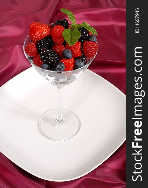 Fresh berries topped with mint in a martini glass resting on a white plate with a wine colored satin background