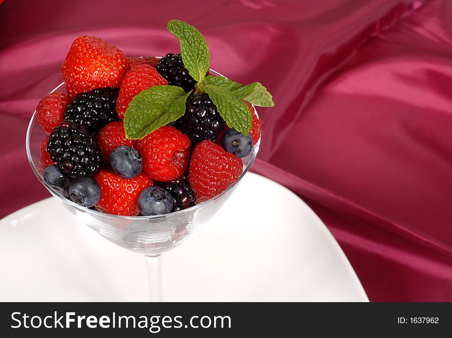 Close up of Fresh berries topped with mint in a martini glass resting on a white plate with a wine colored satin background