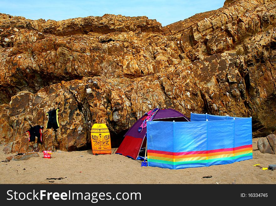 Colourful beach tent and windbreaker with wetsuits, bucket and spade and a body board on a deserted Devon beach in the UK with a lovely rock formation background. Colourful beach tent and windbreaker with wetsuits, bucket and spade and a body board on a deserted Devon beach in the UK with a lovely rock formation background.