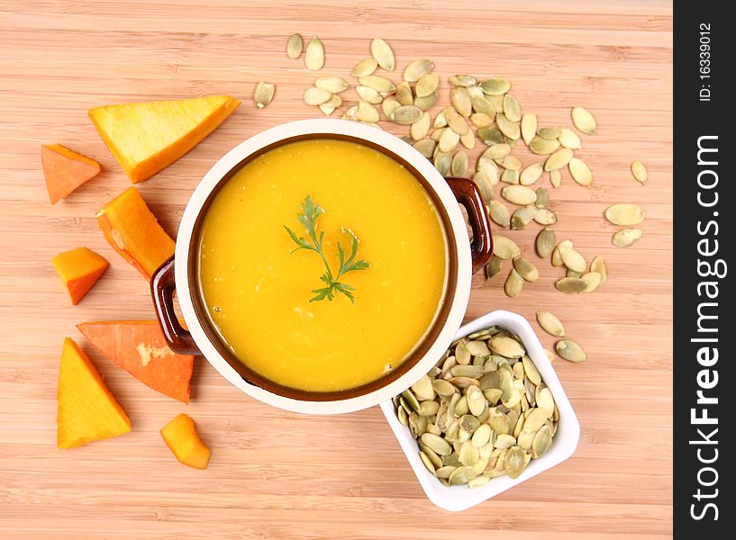 Pumpkin soup decorated with parsley, a bowl of pumpkin seeds and some pieces of pumpkin on wooden background