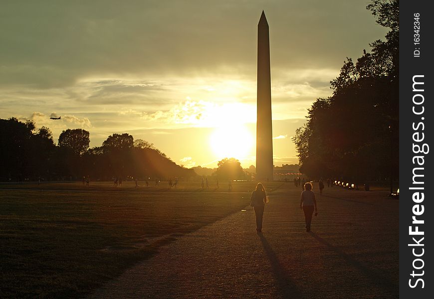 Washington monument at dawn with the flag circle around it and a beautiful sky. two people are walking down the mall towards the monument.