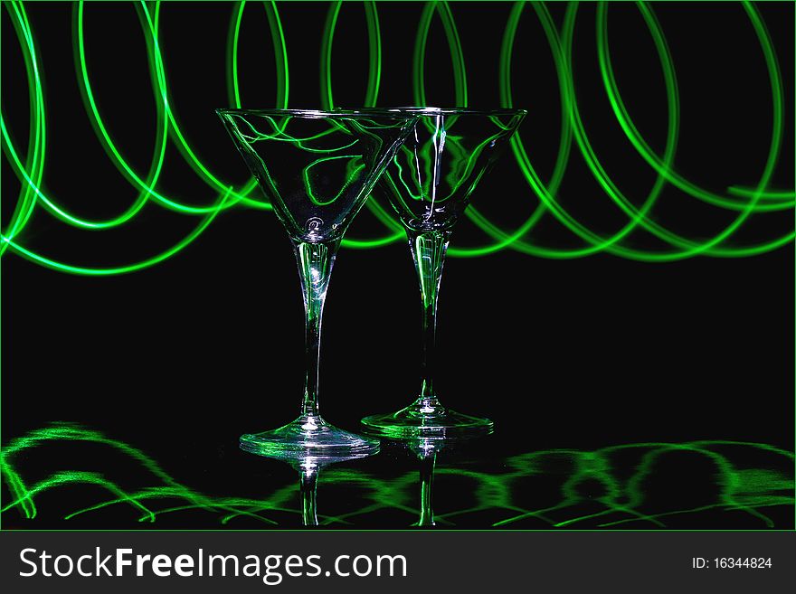 Pair martini glasses with black background and reflective black surface, lit with green led light trails. Pair martini glasses with black background and reflective black surface, lit with green led light trails