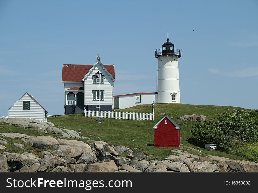 A lighthouse with out-buildings perched on a rocky landscape with green yard and blue, clear sky. A lighthouse with out-buildings perched on a rocky landscape with green yard and blue, clear sky.