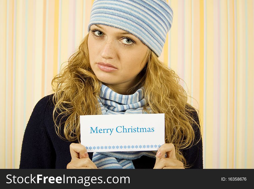 Young beautiful woman in a sweater with a blue hat and scarf holding a card with the text Merry Christmas. Young beautiful woman in a sweater with a blue hat and scarf holding a card with the text Merry Christmas