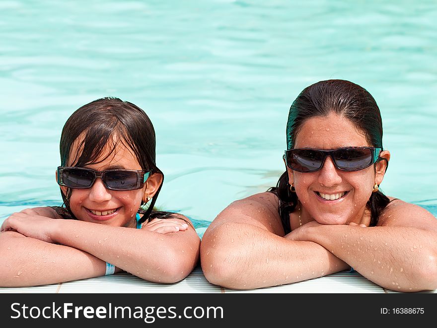 Daughter and mother smiling and wearing sunglasses at the border of a swimming pool. Daughter and mother smiling and wearing sunglasses at the border of a swimming pool