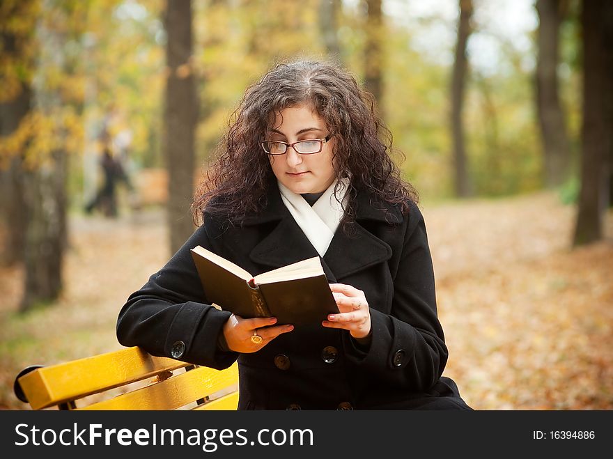 Young woman reading book in autumn park bench