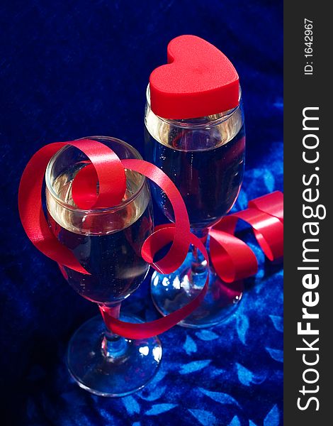 Two glasses of champagne, red twisted ribbon and plastic heart on the blue velvet. Two glasses of champagne, red twisted ribbon and plastic heart on the blue velvet