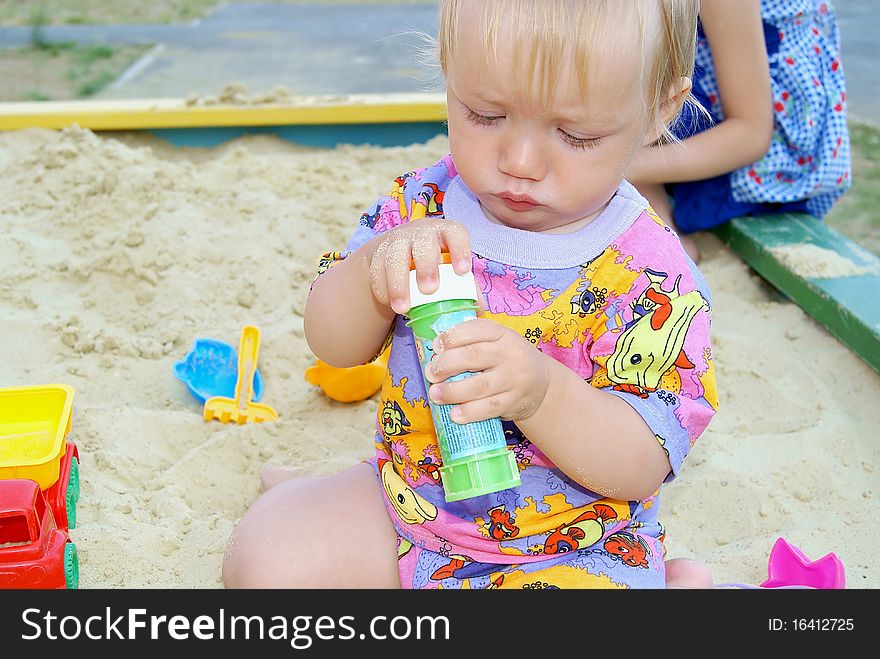 The little boy sits on sand in the summer and opens a cover on a vial. The little boy sits on sand in the summer and opens a cover on a vial