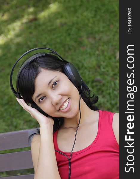 A smiling Asian woman listening to and enjoying the music on the headphone while relaxing in the park. A smiling Asian woman listening to and enjoying the music on the headphone while relaxing in the park