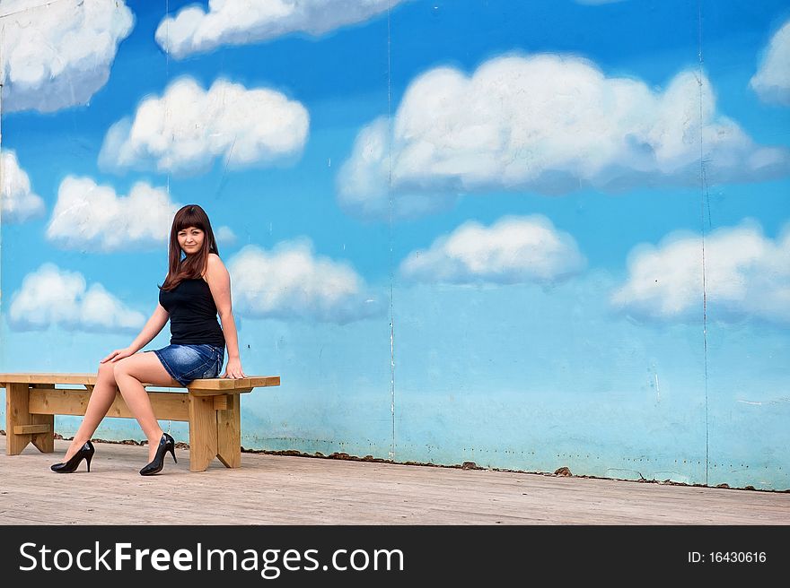Young, woman in short skirt sit on the bench on the background of blue sky drawing. Young, woman in short skirt sit on the bench on the background of blue sky drawing.