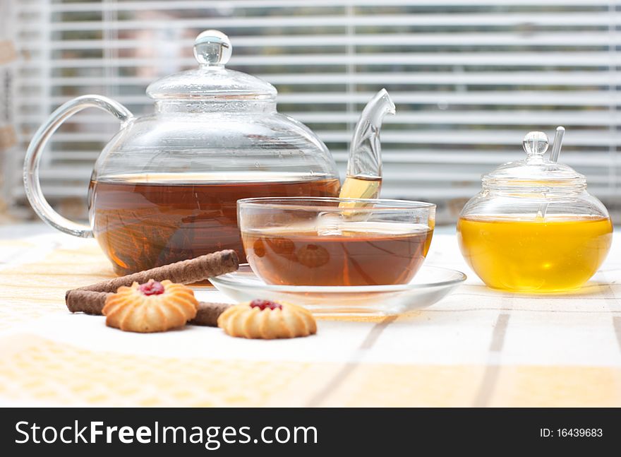 Glass teapot, mug, honey jar and cakes on table near window with jalousie. Glass teapot, mug, honey jar and cakes on table near window with jalousie