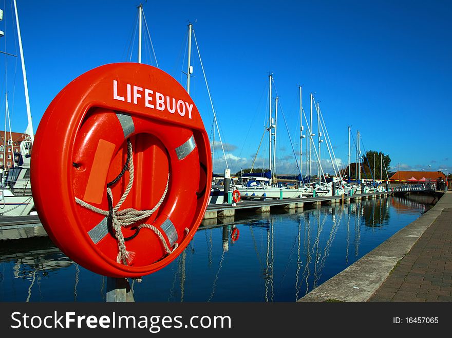 Bright orange lifebuoy captured on a bright sunny day with a deep blue sky on Weymouth Marina, with yachts in the background and reflections in the almost still waters. Bright orange lifebuoy captured on a bright sunny day with a deep blue sky on Weymouth Marina, with yachts in the background and reflections in the almost still waters.