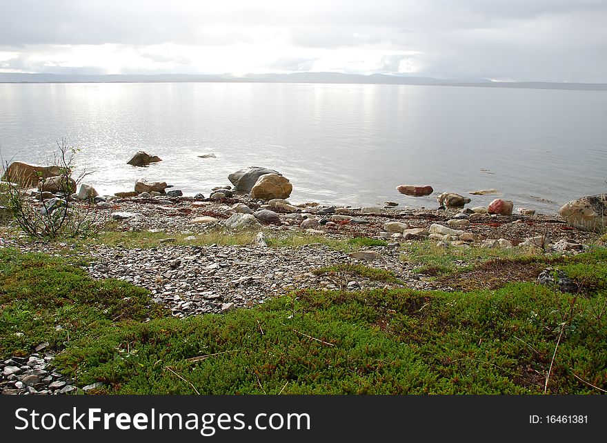 Wild lanscape of Finnmark see cost,Tundra vegetation, stones and transparante water at foreground and sun light crossinf frog and clouds and refrecting in water surfce