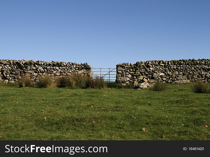 Old metal gate in a dry stone wall in a green grass field and blue sky. Old metal gate in a dry stone wall in a green grass field and blue sky.
