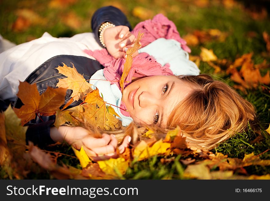 Portrait of a beautiful young blonde girl in a pink scarf lying on green grass among the autumn maple leaves on a bright sunny day. Portrait of a beautiful young blonde girl in a pink scarf lying on green grass among the autumn maple leaves on a bright sunny day