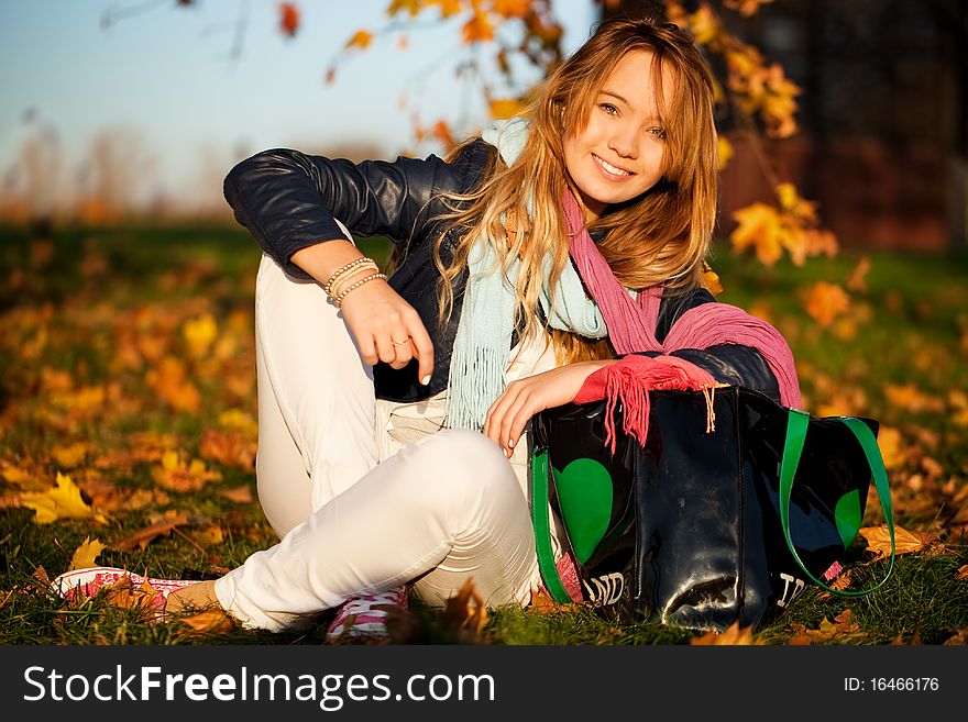 Portrait of a beautiful young blond smiling girl sitting in the park grass among autumn maple leaves on a bright sunny day. Portrait of a beautiful young blond smiling girl sitting in the park grass among autumn maple leaves on a bright sunny day